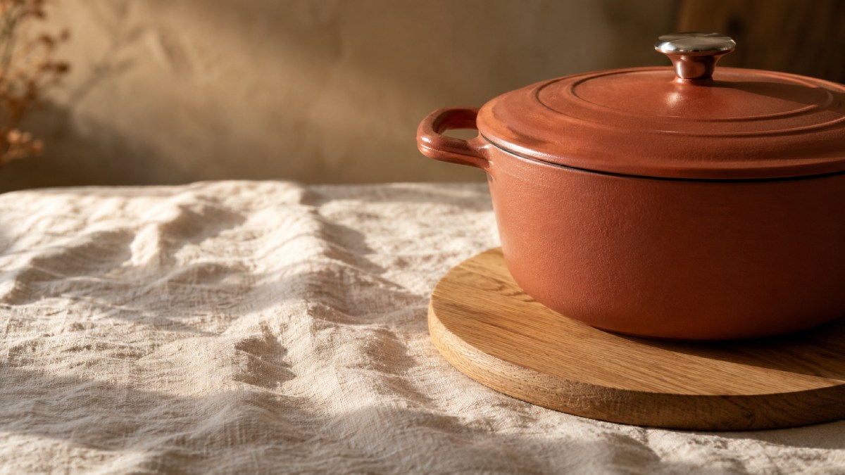Cocotte en fonte terracotta posée sur un sous-plat rond en chêne, sur une nappe en lin lavé écru, en lumière rasante