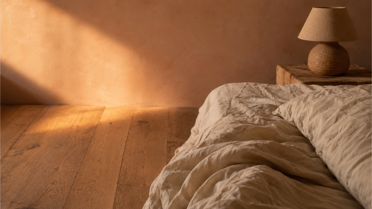 Coin d'un lit en lin lavé écru froissé et sol en chêne brut, éclairés par une lumière rasante de fin de journée, dans une chambre aux murs beige rosé