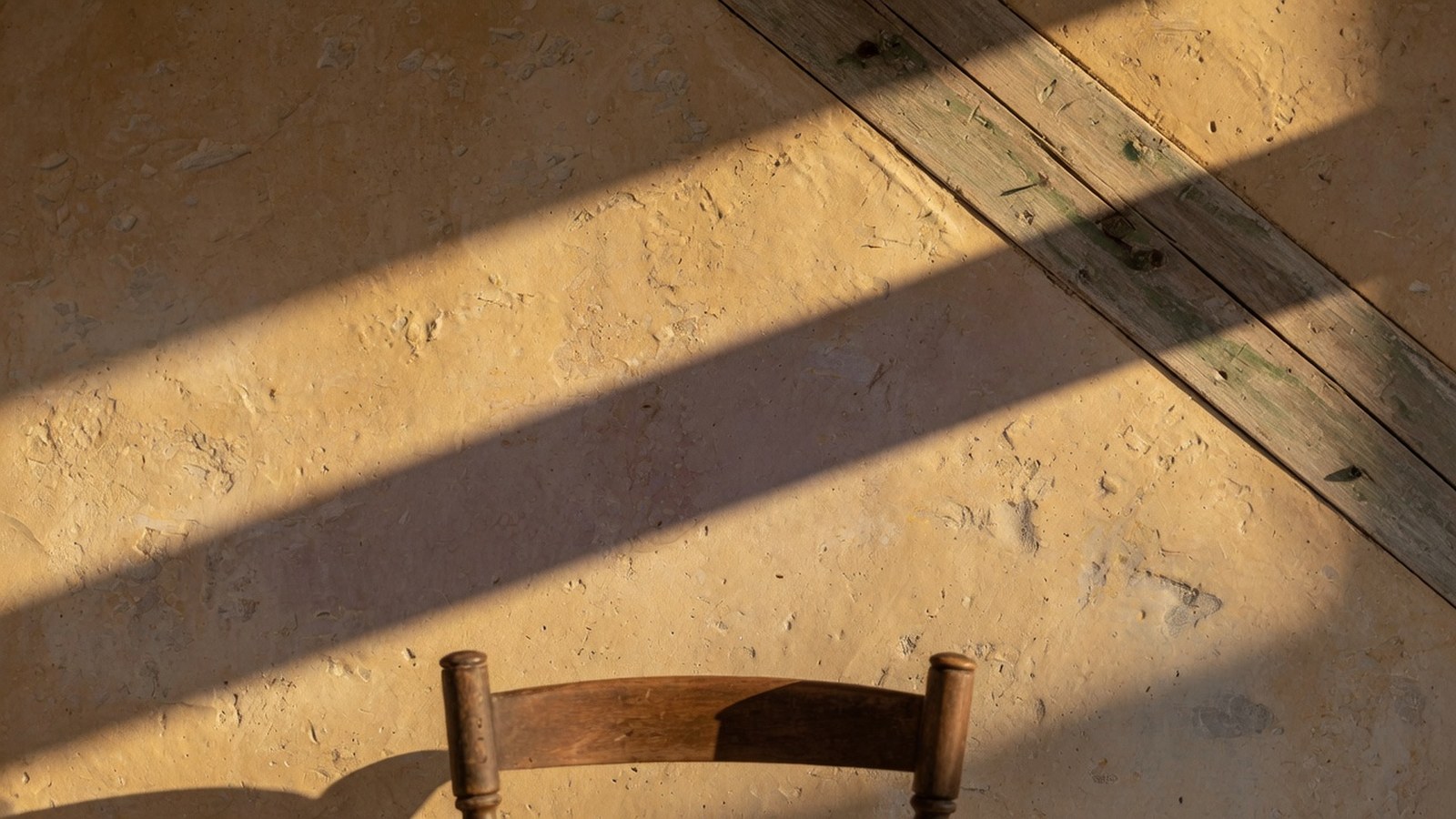Mur chaulé ocre avec badigeon texturé inégal, poutre bois vieillie en diagonale, lumière rasante