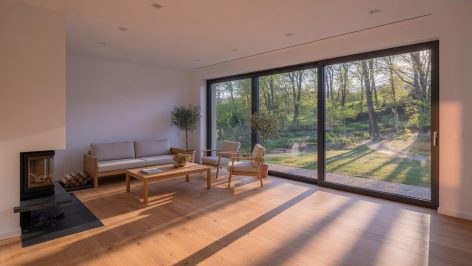 Salon d'une maison passive neuve au Luxembourg avec baies vitrées pleine hauteur, parquet chêne clair et lumière de fin d'après-midi sur jardin boisé