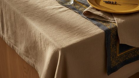 Coin de table en chêne recouvert d'une nappe en lin froissé beige, avec un chemin de table en indigo et jaune posé en diagona
