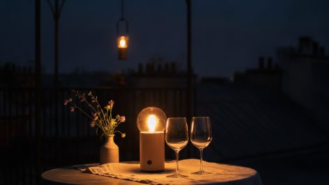 Lampe sans fil à globe ambré éclairant deux verres à vin sur une table en zinc, nappe en lin froissé, terrasse en soirée.