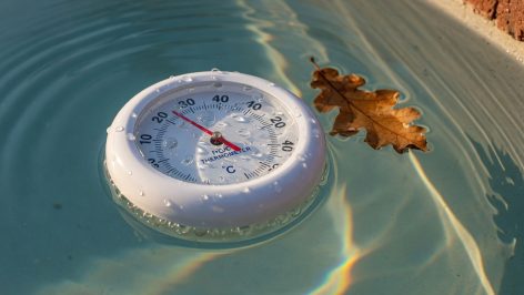 Thermomètre flottant sur piscine bleu-vert : gouttelettes d'eau, feuille morte et ondulations, lumière automnale.