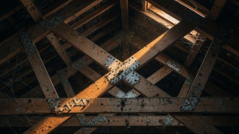 Vue en plongée de la charpente en W d'un comble des années 70, poutres bois brutes et connecteurs métalliques éclairés par un